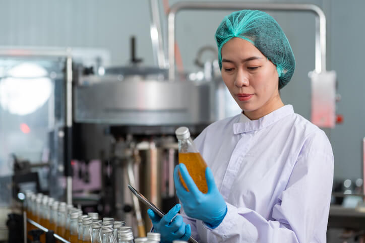 A female product development technologist checking product bottles of fruit juice on the conveyor belt in the beverage factory after completing her food safety training